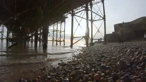 Underneath-Brighton-Pier,-pebble-beach-with-sun-flare,-seagulls-flying-past-and-waves-lapping-at-the-shore