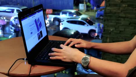 Male-person-surfing-the-net-and-performing-online-work-on-his-laptop-computer-at-a-coffee-shop,-with-heavy-vehicular-traffic-showing-through-a-glass-wall