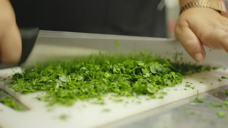 Woman-hands-cutting-green-parsley-in-a-restaurant-kitchen-with-a-professional-knife-over-a-white-plastic-chopping-board