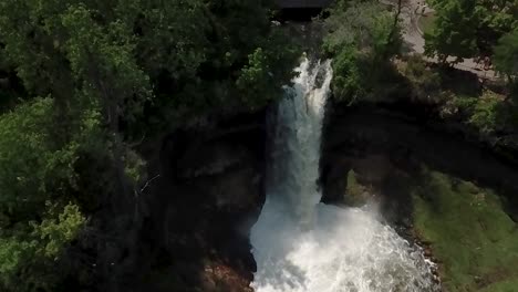 Wide-aerial-shot-in-slow-motion-looking-down-and-flying-over-the-Minnehaha-waterfall-with-its-white,-rushing-water-and-then-flying-over-the-green-tree-tops-with-trails-below