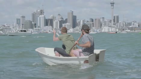 Zwei-Ambitionierte-Segler-Paddeln-Ein-Schlauchboot-Gegen-Die-Strömung-Zu-Ihrem-Boot-Mit-Auckland-City-Im-Hintergrund