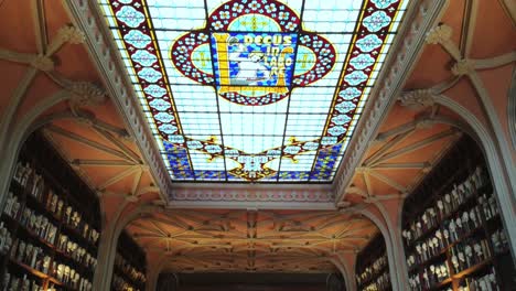 Tilt-up-from-people-posing-on-iconic-Livraria-Lello-staircase-in-Porto,-Portugal-to-ornate-stained-glass-ceiling