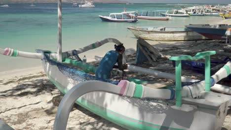 Asiatischer-Mann-Sitzt-Auf-Einem-Boot-An-Einem-Sandstrand,-Macht-Eine-Pause-Und-Schaut-Auf-Sein-Smartphone-Auf-Gili-Air,-Indonesien