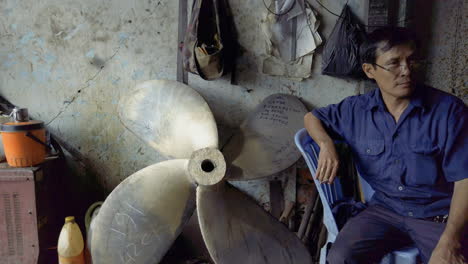 Hombre-Vietnamita-Fumando-Un-Cigarrillo-Dentro-De-Un-Taller-Junto-Al-Río-En-La-Ciudad-De-Ho-Chi-Minh-En-Vietnam,-Donde-Se-Fabrican-Hélices-Para-Grandes-Barcos-Fluviales