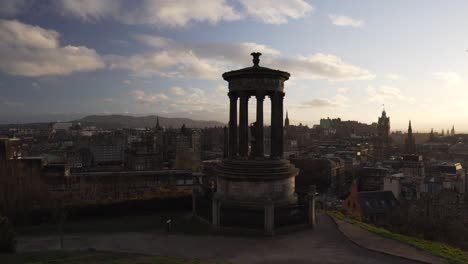 Tracking-follow-shot-from-Calton-hill-with-people-walking-and-Nelson-monument-in-the-foreground-with-nice-sunset-light-and-clouds-overlooking-the-city-of-Edinburgh,-Scotland