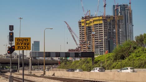 Daily-commute-timelapse-as-vehicles-and-trains-pass-by-a-tall-building-under-construction-in-Tel-Aviv,-Israel