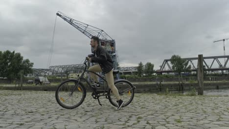 Back-view-of-young-male-in-casual-wear-taking-bike-parked-on-urban-waterfront-near-industrial-crane-and-moving-away