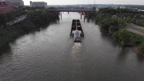 Tugboat-pushes-and-guides-large-commercial-transport-barge-down-river-towards-bridge-by-downtown-Nashville-city,-Illinois,-rising-aerial-pull-back