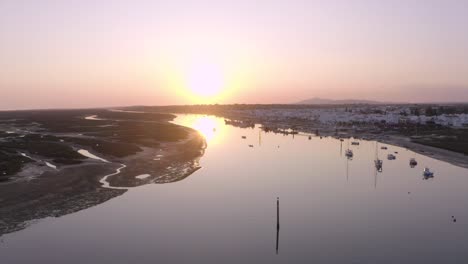 Drone-Vuela-Hacia-Una-Espectacular-Puesta-De-Sol-Sobre-Barcos-Y-Un-Río-Bordeado-De-Casas,-Muelles-Y-Muelles,-Luz-Tavira,-Antena