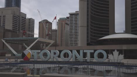 The-Famous-Toronto-Sign-And-Canadian-Flag-At-The-Nathan-Phillips-Square-In-Toronto,-Canada-With-Downtown-Skyline-In-The-Background---midshot