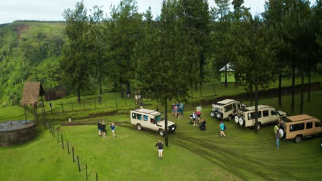 Safari-Jeep-Geländewagen-Mit-Touristen,-Die-Auf-Einer-Schlammigen-Straße-Im-Dschungelwald-Im-Ländlichen-Tansania-Fahren,-An-Einem-Tor-Vorbei-Und-Auf-Einem-Grasparkplatz-In-Einem-Buschdorf-Ankommen,-Drohnenflugaufnahme-4k