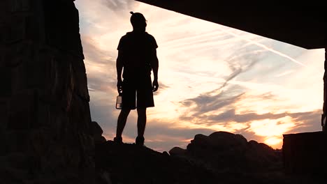 Silhouette-Eines-Rucksacktouristen-Außerhalb-Einer-Höhle,-Der-Den-Orangefarbenen-Horizont-Betrachtet