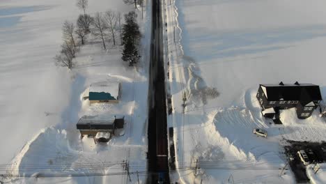 Eine-4K-Drohnenaufnahme-Schwenkt-Von-Einer-Verschneiten-Straße-Und-Einem-Dorf-Nach-Oben-Und-Zeigt-Im-Winter-Bei-Sonnenuntergang-Und-Zur-Goldenen-Stunde-Die-Schneebedeckten-Berge-Des-Skigebiets-Niseko-In-Japan