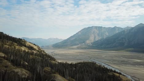 Glacial-river-running-through-the-valley-with-mountains,-forest,-and-trees-in-the-background