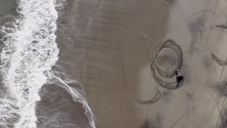 Dirt-Bike-rider-preforming-Donuts-on-a-sandy-beach-in-Bali,-with-calm-waves-breaking,-Top-down-aerial-view