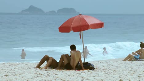 Dos-Chicos-Relajándose-En-La-Arena-De-La-Playa-De-Ipanema-En-Un-Día-Increíble-Con-Gente-Jugando-En-El-Fondo