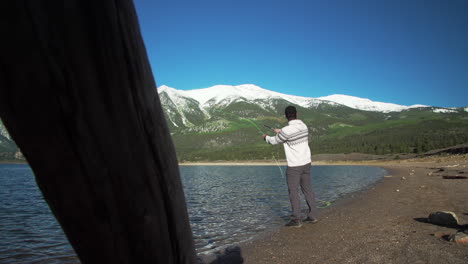 Wide-dolly-shot-of-a-fly-fisherman-casting-his-lure-into-a-deep-blue-lake-in-front-of-gorgeous,-snow-capped-mountains