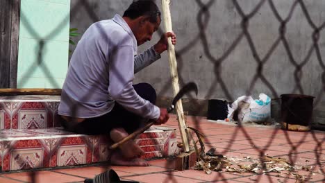 Old-Vietnamese-man-remove-bark-from-wooden-stick-with-sickle-in-front-of-house