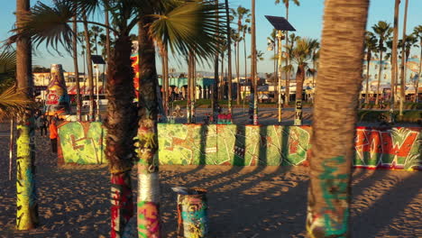 Drone-shot-of-venice-beach-boardwalk-during-sunset-showing-palm-trees,-graffiti-walls,-skateboarding-and-people-taking-photos