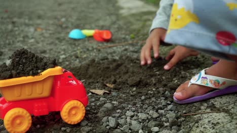 Nahaufnahme-Der-Hände-Eines-Glücklichen-Kindes,-Das-Auf-Der-Straße-Mit-Steinen,-Sand-Und-LKW-Spielzeug-Spielt