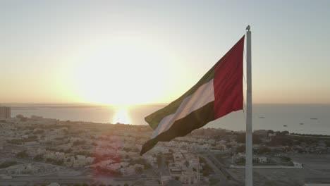 Vista-Aérea:-Bandera-De-Los-Emiratos-árabes-Unidos-Ondeando-En-El-Viento,-El-Cielo-Y-El-Sol-De-Fondo-El-Símbolo-Nacional-De-Los-Emiratos-árabes-Unidos