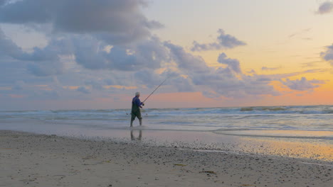 Old-man-is-fishing-on-the-beach-throwing-bait-with-fishing-rod-in-the-surf-during-sunset