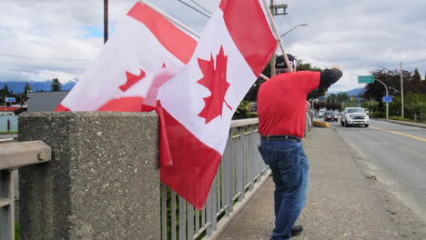 Ciudadano-Canadiense-Patriótico-Sosteniendo-Banderas-De-Canadá-En-La-Calle,-Alegre-Saludando-A-Los-Autos-Que-Pasan