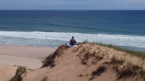 Drone-Pullback-View-Tourist-Sitting-on-Large-Sand-Dune-Waves-to-People-on-Beach