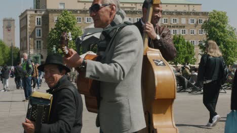 Three-street-musicians-talking-to-people-passing-by-and-playing-instruments-in-downtown-Stuttgart-square-at-noon,-Germany,-Europe,-panning-view-angles