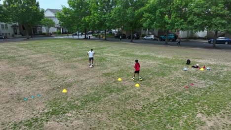 Aerial-orbit-shot-of-father-training-soccer-with-son-on-grass-field-in-park-of-american-town