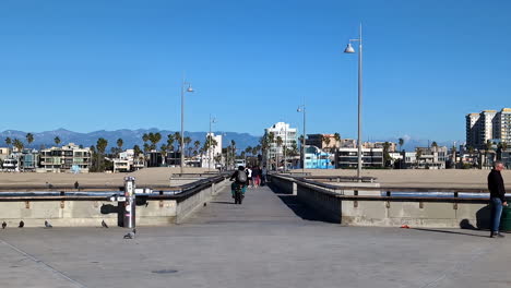 Gente-Disfrutando-De-Un-Día-Soleado-En-El-Muelle-De-Venice-Beach-En-Los-Ángeles-Con-Montañas-Al-Fondo