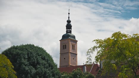 Tower-of-Varaždin-Cathedral-of-the-Assumption-of-the-Blessed-Virgin-Mary,-rises-above-the-lush-greenery-and-rooftops,-set-against-a-partly-cloudy-sky