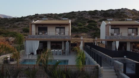 Slow-drone-flight-next-to-two-houses-with-outdoor-pool-at-sunrise-in-Greece-on-the-island-of-Crete---You-can-see-palm-trees-and-rocky-mountains