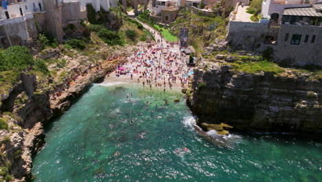 Lama-Monachile-Beach-With-Crowd-Of-People-In-Polignano-a-Mare,-Italy---Aerial-Pullback