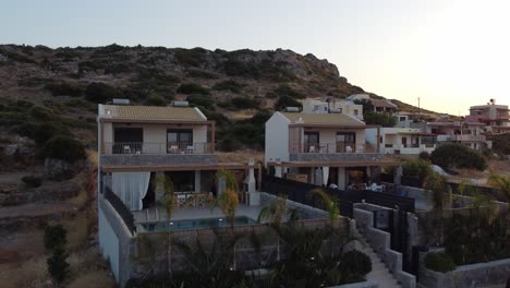 Drone-flying-slowly-near-two-houses-with-outdoor-pool-at-sunrise-in-Greece-on-the-island-of-Crete---You-can-see-palm-trees-and-rocky-mountains