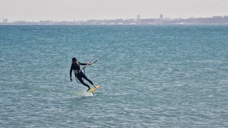 Mann-Beim-Kitesurfen-Mit-Einem-Tragflächenboot-An-Einem-Windigen-Tag-Mit-Dem-Industriehafen-Im-Hintergrund