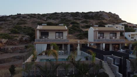 Drone-flies-fast-in-the-air-near-two-houses-with-outdoor-pool-at-sunrise-in-Greece-on-the-island-of-Crete---you-can-see-palm-trees-and-rocky-mountains