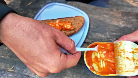 Closeup-of-person-using-plastic-fork-scooping-sardines-in-sauce-from-can-onto-wooden-picnic-bench