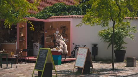 Puesto-En-El-Mercado-De-Artesanía-Tradicional-De-Varazdin,-Con-Un-Gran-Osito-De-Peluche-En-Exhibición,-Carteles-De-Pizarra-Con-Listados-De-Productos-Y-Una-Bicicleta-Al-Fondo.