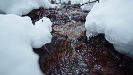 Snowy-stream-flows-between-rocks-and-branches-in-a-serene-winter-forest