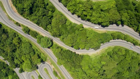 A-winding-mountain-road-surrounded-by-lush-green-trees-on-a-sunny-day,-aerial-view