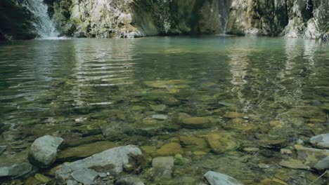 A-continuous-cascade-of-water-from-a-distant-waterfall-feeds-into-a-serene-pond,-its-surface-shimmering-with-hues-of-green