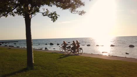 Three-women-ride-bikes-by-the-sea-at-sunset-at-Gotland,-Sweden,-slomo