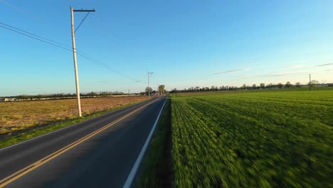 FPV-drone-flight-over-rural-road-surrounded-by-farm-fields-during-golden-sunset