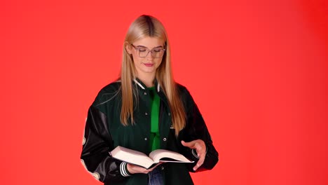 Pretty-Blonde-Student-Standing-Reading-a-Book-Against-Red-Background,-Studio-Shot