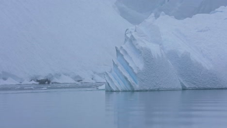 Huge-Iceberg-Floating-in-Cold-Ocean-Water-by-Snow-Capped-Coast-of-Antarctica