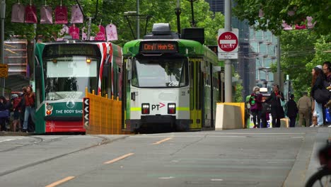 Trams-glide-along-tree-lined-street-with-commuters-disembarking-and-boarding-the-tram-during-rush-hour-in-Melbourne-city's-bustling-Central-Business-District,-swarm-of-pedestrians-crossing-the-road