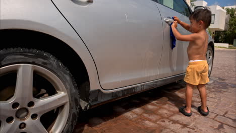 Zeitlupe-Eines-Jungen-Mexikanischen-Lateinamerikanischen-Jungen-Ohne-Hemd,-Der-An-Einem-Sonnigen-Tag-Das-Familienauto-Mit-Einem-Nassen-Blauen-Lappen-Putzt