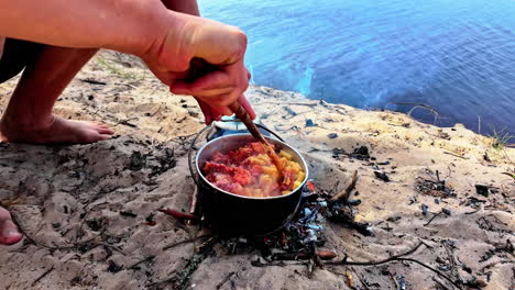 Barefoot-man-squats-on-sand-cooking-fresh-food-over-wooden-fire-on-beach-making-pasta-with-sauce,-slow-motion