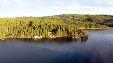 Aerial-view-of-a-serene-lake-surrounded-by-a-dense-Norwegian-forest-with-scattered-cabins-along-the-shoreline,-bathed-in-soft-sunlight